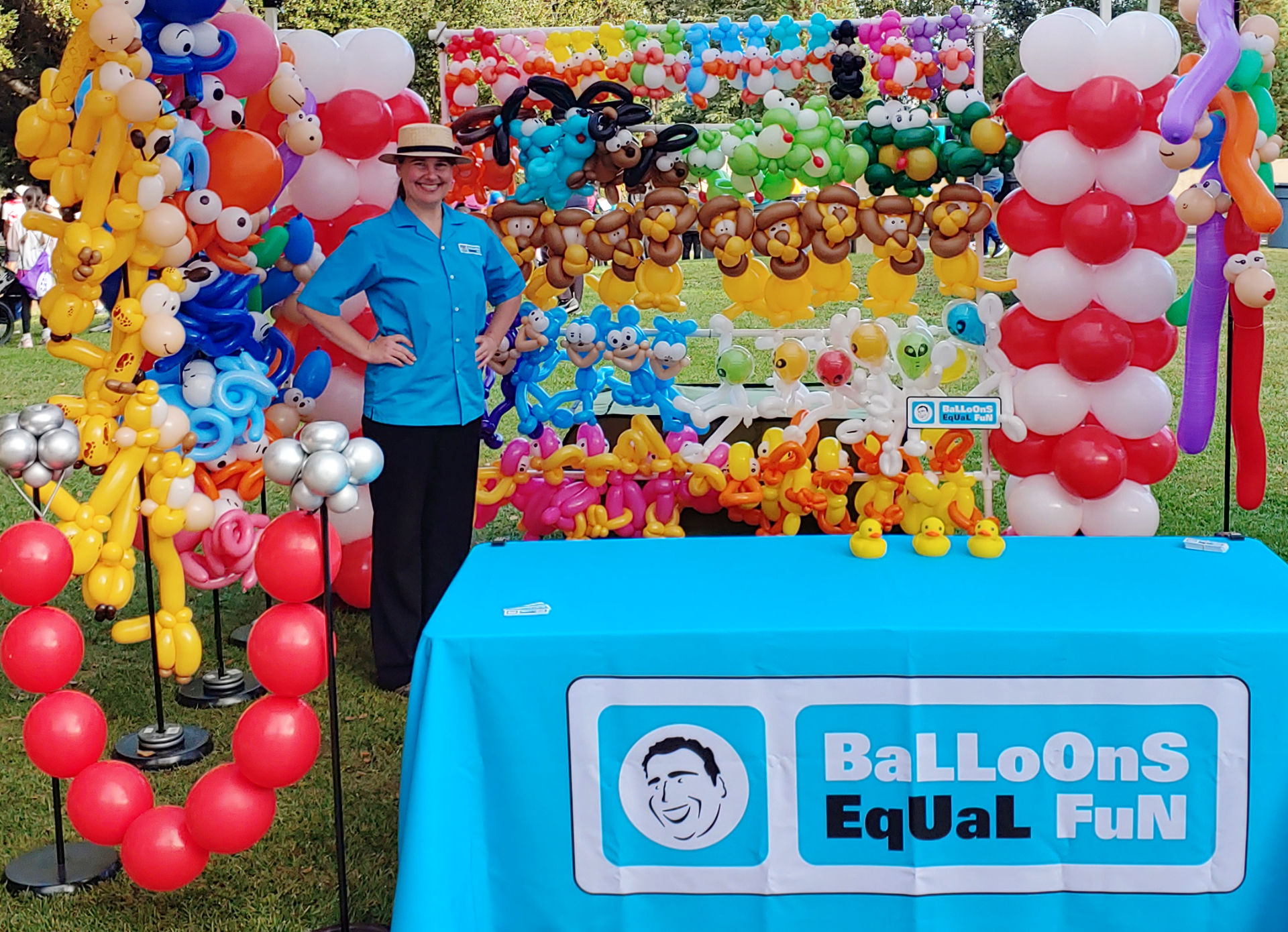 Carnival booth featuring Jessica! She is wearing a Balloons Equal Fun uniform shirt and a carnival barker hat. There are 3 rubber duckies on the table for the 'Find the Squeaking Duckie!' game.