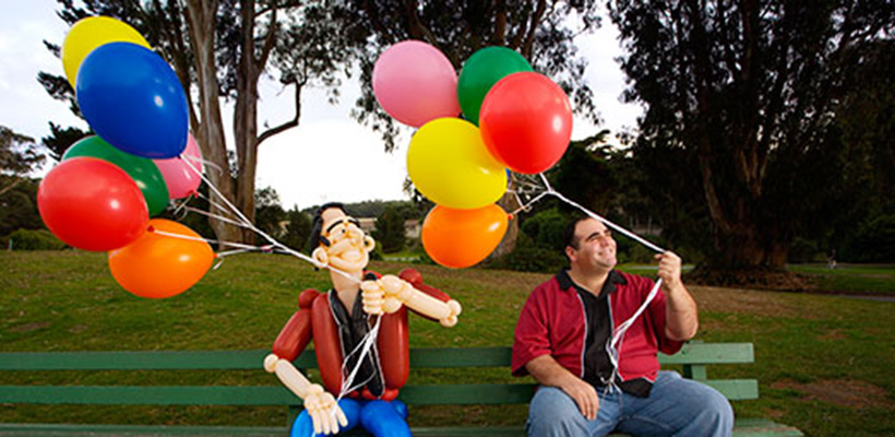 Brian on a green park bench holding a bunch of helium balloons. There is a life size balloon version of Brian seated next to him, holding a matching bunch of helium balloons.